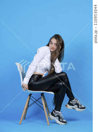 Pretty caucasian woman in white blouse, resting head on hand, while sitting on chair cross legged. Side view of cute girl smiling, while posing at camera, isolated on blue. Concept of female posing. 110953044
