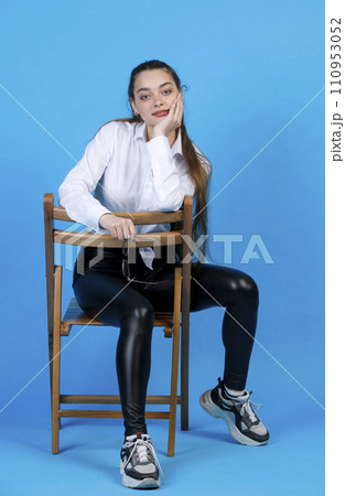 Young girl with ponytail resting head on hand, while sitting on wooden chair backwards. Front view of cute lady posing, while looking at camera, isolated on blue background. Concept of posing. 110953052