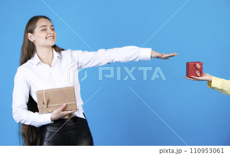 Surprised, pretty birthday girl in elegant shirt, with closed eyes, while holding out arm to receiving wrapped small gift box from unrecognizable male hand, isolated on blue. Concept of expectation. Surprised, pretty birthday girl in elegant shirt, with closed eyes, while holding out arm to receiving wrapped small gift box from unrecognizable male hand, isolated on blue. Concept of expectation. 110953061
