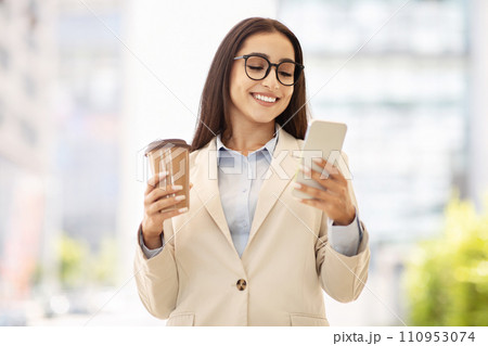 Content businesswoman in a stylish beige jacket holds a coffee cup and smartphone, engaged in reading 110953074