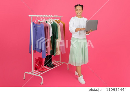 Online Stylist. Happy asian woman holding laptop while standing near clothing rack 110953444
