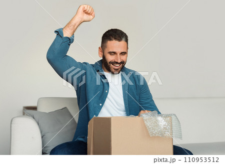 Joyful middle aged guy unpacking carton box shaking fist indoors Joyful middle aged guy unpacking carton box shaking fist indoors 110953512