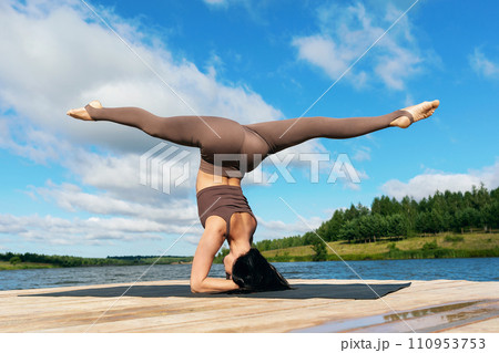 Attractive woman performing Shirsasana exercise with Hanumanasana, inverted asana, headstand with longitudinal splits, training on the shore of a lake on a warm sunny morning 110953753