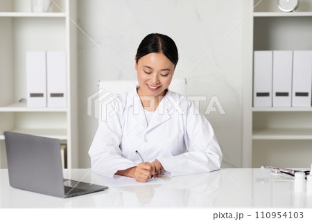 Smiling Asian Woman Doctor Writing Notes While Sitting At Desk In Clinic Smiling Asian Woman Doctor Writing Notes While Sitting At Desk In Clinic 110954103