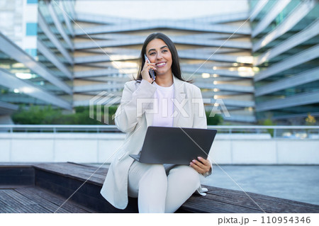 businesswoman communicating talking on cellphone using her laptop in city businesswoman communicating talking on cellphone using her laptop in city 110954346