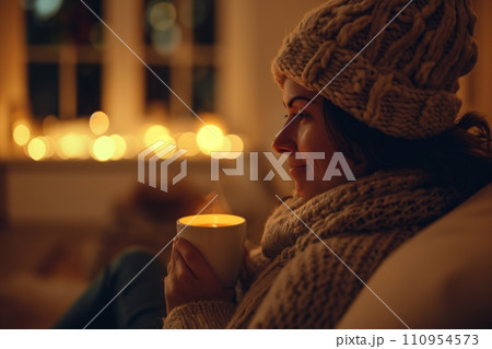 A cozy and comforting scene of a woman in a woolen hat, wrapped in a warm blanket, sitting in a living room and sipping tea, the ambiance is homely and relaxing 110954573