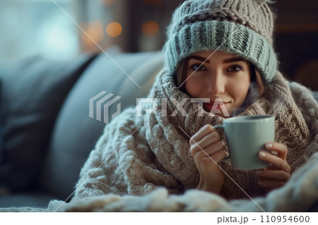 A woman in a cozy woolen hat, wrapped in a plush blanket, holding a steaming cup of tea, set in a comfortable and inviting living room 110954600