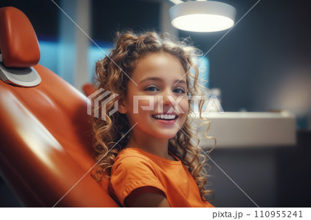 A young girl with curly hair is sitting in a dental chair and smiling at the camera. 110955241