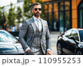 A man in a grey suit and sunglasses stands in front of two cars. A man in a grey suit and sunglasses stands in front of two cars. 110955253