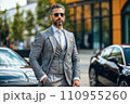 A man in a grey suit and sunglasses stands in front of two cars. A man in a grey suit and sunglasses stands in front of two cars. 110955260