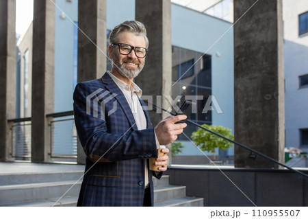 Smiling senior professional man in business suit using smartphone outside contemporary office building. 110955507