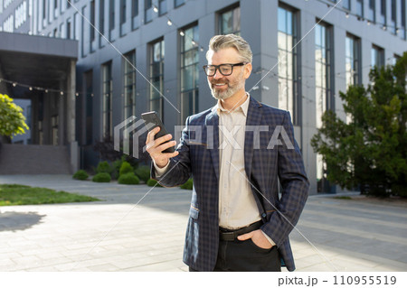 Confident mature male executive using a mobile phone, standing outside a contemporary corporate office setting. 110955519