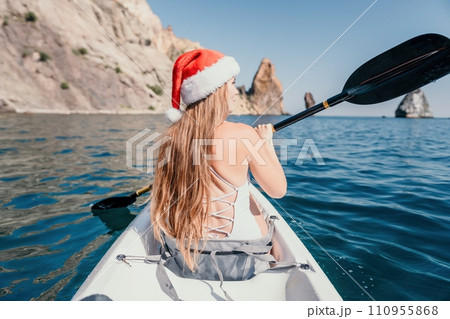 Woman in kayak back view. Happy young woman in Santa hat floating in kayak on calm sea. Summer holiday vacation and cheerful female people relaxing having fun on the boat. 110955868