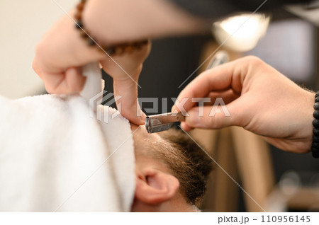 A barber shaves the cheek of a bearded customer with dangerous razor. Shaving the contour of the beard for the correct shape. 110956145