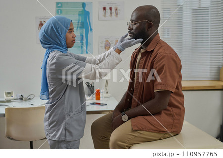 Young muslim doctor in uniform and protective gloves touching lymph nodes of her black male patient Young muslim doctor in uniform and protective gloves touching lymph nodes of her black male patient 110957765
