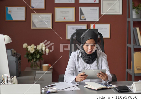 Young muslim doctor sitting at her desk and browsing digital tablet taking break from paperwork 110957836