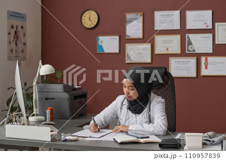 Young female doctor in hijab sitting at clinic desk and attentively filling documents 110957839