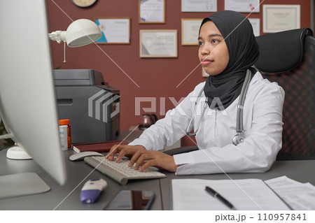 Young female doctor in hijab sitting in hospital office and typing on desktop computer 110957841