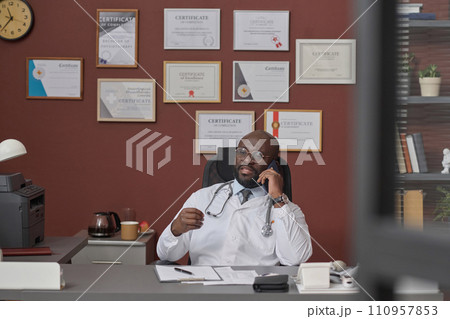 African american doctor talking on his smartphone while sitting at his workplace in clinic 110957853