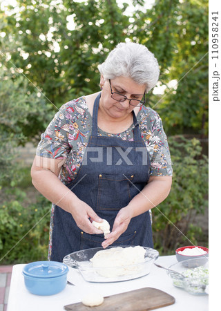 Front view of old woman wearing glasses and apron, looking down, holding dough, rolling buns outdoor. Concept of baking. Front view of old woman wearing glasses and apron, looking down, holding dough, rolling buns outdoor. Concept of baking. 110958241