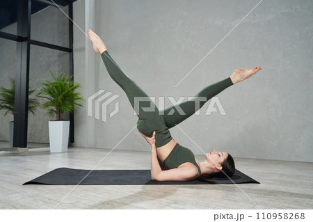 Young slender woman in tight leggings practicing yoga legs up pose on yoga mat at home. Side view of focused girl doing hatha yoga postures in modern indoor space. Concept of yoga practice, sport.  110958268