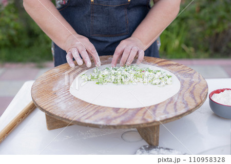 Close up of old female cook, chef wearing apron making pita, putting cheese filling on dough. Concept of fast food. Close up of old female cook, chef wearing apron making pita, putting cheese filling on dough. Concept of fast food. 110958328