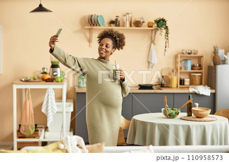 Medium long shot of happy pregnant African American woman taking selfie with glass of green smoothie in spacious kitchen 110958573