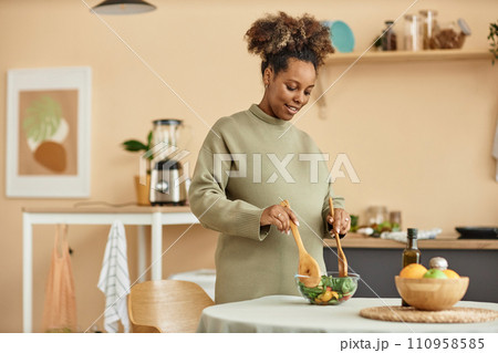 Waist up shot of smiling pregnant African American woman mixing fresh salad in bowl standing at kitchen table 110958585