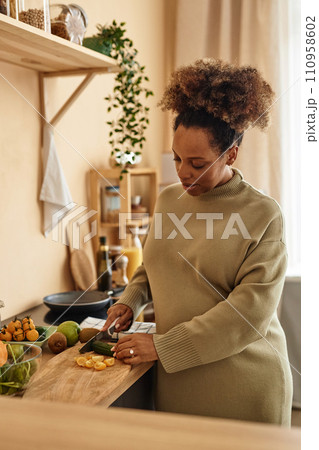 Vertical shot of pregnant Black woman wearing pastel green sweater dress cutting vegetables for salad on wooden board in kitchen Vertical shot of pregnant Black woman wearing pastel green sweater dress cutting vegetables for salad on wooden board in kitchen 110958602