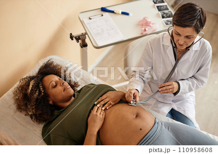 Top down view of pregnant Black woman lying on examination table in medical center exposing big belly while senior doctor listening to baby heart sounds 110958608