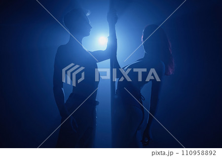 Silhouettes of woman and man dancing on stage holding on to pole illuminated with blue spotlight beam Silhouettes of woman and man dancing on stage holding on to pole illuminated with blue spotlight beam 110958892
