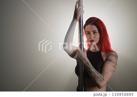 Waist up shot of red-haired young Caucasian woman with arm tattoos looking at camera and holding on to pole standing in studio backlight 110958895