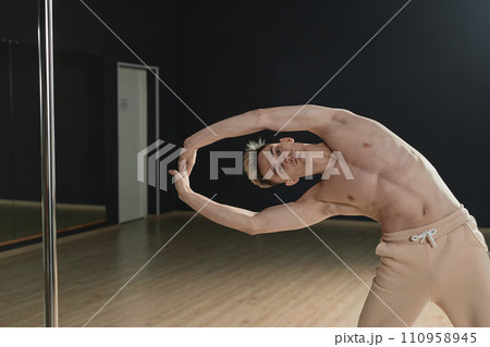 Medium long shot of young Caucasian male pole dancer doing stretching exercise standing in studio Medium long shot of young Caucasian male pole dancer doing stretching exercise standing in studio 110958945