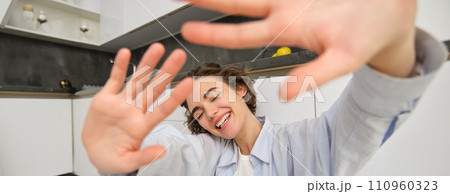 Lifestyle and people concept. Young happy woman hides from camera, extends hands forward, laughs and smiles carefree, spends time at home with joy Lifestyle and people concept. Young happy woman hides from camera, extends hands forward, laughs and smiles carefree, spends time at home with joy 110960323