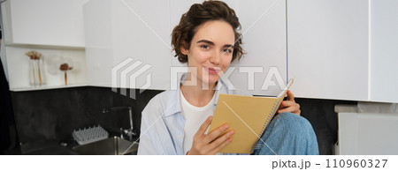 Portrait of brunette girl with short hair, holds planner, reads journal book on kitchen counter, enjoys leisure time at home Portrait of brunette girl with short hair, holds planner, reads journal book on kitchen counter, enjoys leisure time at home 110960327