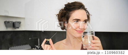 Portrait of sportswoman drinking water, taking vitamins, dietry supplements for healthy skin, having omega-3 pill, standing in her kitchen in workout clothing 110960382