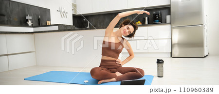 Young woman looking at digital tablet, connects to stretching fitness online class, workout, sits on yoga rubber mat at home, follows gym instructor exercises remotely Young woman looking at digital tablet, connects to stretching fitness online class, workout, sits on yoga rubber mat at home, follows gym instructor exercises remotely 110960389