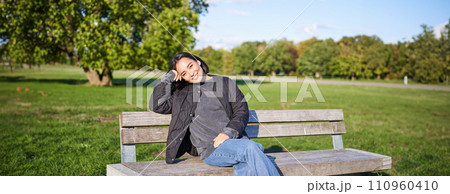 Smiling young asian woman in outdoor jacket, sitting on bench in green sunny park, resting alone, relaxing on fresh air Smiling young asian woman in outdoor jacket, sitting on bench in green sunny park, resting alone, relaxing on fresh air 110960410