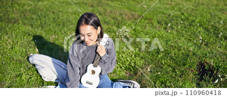 Smiling asian girl learns how to play ukulele on laptop, video chat with music teacher, sitting with instrument in park on grass Smiling asian girl learns how to play ukulele on laptop, video chat with music teacher, sitting with instrument in park on grass 110960418