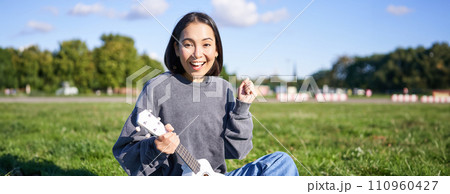 Portrait of excited asian girl, sitting with ukulele in park on green grass, looking amazed and surprised with good news Portrait of excited asian girl, sitting with ukulele in park on green grass, looking amazed and surprised with good news 110960427