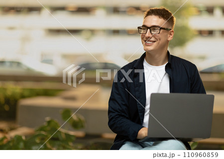 Smiling young european man in casual, glasses, enjoy lifestyle, work on laptop, sit outdoor 110960859
