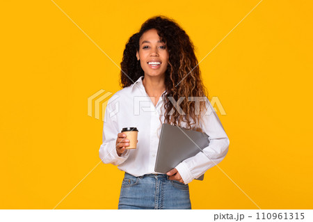Happy black woman holding laptop and coffee on yellow background Happy black woman holding laptop and coffee on yellow background 110961315