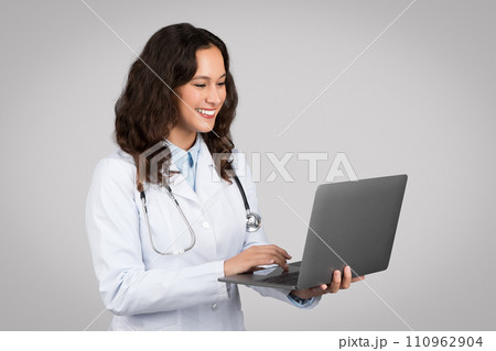 Happy caucasian lady doctor therapist in white coat typing on laptop, isolated on gray background studio shot 110962904