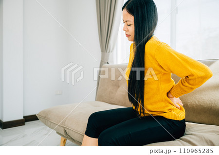 A woman in Asia suffering unbearable lower back pain while seated on a sofa. Depicting chronic backache discomfort and the necessity of medical care for back problems. healthcare and problem concept 110965285