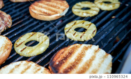 Grilled hamburger buns with pineapple on stainless BBQ grill with flames on dark background. Food and cuisine concept. Grilled hamburger buns with pineapple on stainless BBQ grill with flames on dark background. Food and cuisine concept. 110965324