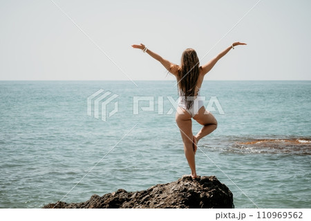 Woman sea yoga. Back view of free calm happy satisfied woman with long hair standing on top rock with yoga position against of sky by the sea. Healthy lifestyle outdoors in nature, fitness concept. Woman sea yoga. Back view of free calm happy satisfied woman with long hair standing on top rock with yoga position against of sky by the sea. Healthy lifestyle outdoors in nature, fitness concept. 110969562