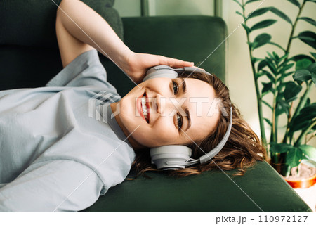 Joyful young woman wearing wireless headphones listening to music while lying on the sofa, enjoying Joyful young woman wearing wireless headphones listening to music while lying on the sofa, enjoying 110972127