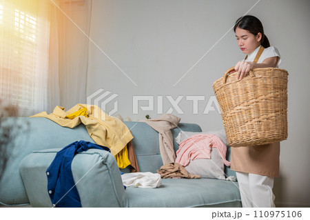 Asian housewife woman holding basket with heap of different clothes on sofa in living room, Beautiful housekeeper doing housework holds wooden basket of clean clothes to washing at home Asian housewife woman holding basket with heap of different clothes on sofa in living room, Beautiful housekeeper doing housework holds wooden basket of clean clothes to washing at home 110975106
