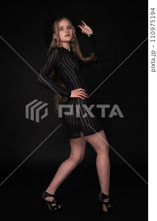 Blonde fashion model looks amazing in black short knitted translucent dress, wide-brimmed felt hat, high heels. Full length portrait captures her from three-quarter rear view against black background 110975194