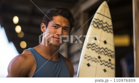Happy Asian man standing on the beach with a surfboard on a bright summer day. Smiling muscular young Asian guy posing on the shore with a board. Portrait of a fit Chinese surfer on a vacation. 110976809
